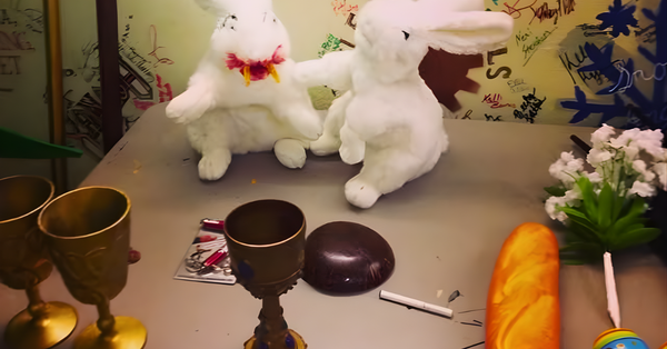 A theatre props table for a production of Monty Python's Spamalot musical. Two white plush rabbits sit on a table surrounded by gold chalices, a baguette, a chocolate egg, flowers, and a scribbled wall, creating a whimsical scene.